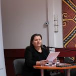 Kobieta wygłasza referat. W rękach trzyma kartki. W tle widać roll-up Polskiego Seminarium Etnomuzykologicznego.