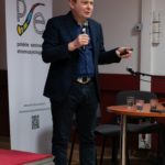 Mężczyzna stoi i mówi do mikrofonu. W tle widać roll-up Polskiego Seminarium Etnomuzykologicznego.