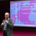 Mężczyzna stoi i mówi do mikrofonu. W tle ekran z wyświetlonym plakatem konferencji.