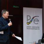Mężczyzna mówi do mikrofonu. Kobieta siedzi i słucha. W tle roll-up Polskiego Seminarium Etnomuzykologicznego.