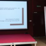 Kobieta siedzi i wygłasza referat. W tle widać roll-up Polskiego Seminarium Etnomuzykologicznego. Na ekranie wyświetlana jest prezentacja.
