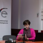 Kobieta siedzi i wygłasza referat. W tle widać roll-up Polskiego Seminarium Etnomuzykologicznego.