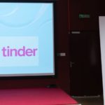 Kobieta siedzi i wygłasza referat. W tle widać roll-up Polskiego Seminarium Etnomuzykologicznego oraz prezentację wyświetlaną na ekranie.