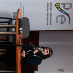 Mężczyzna siedzi i wygłasza referat. W ręku trzyma mikrofon. W tle widać roll-up Polskiego Seminarium Etnomuzykologicznego.