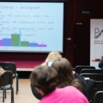 Grupa osób słucha wykładu. Na ekranie wyświetlana jest prezentacja multimedialna.