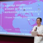 Kobieta stoi i wygłasza referat. W rękach trzyma kartki i mikrofon. W tle na ekranie wyświetlony jest plakat konferencji.