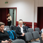 Uczestnicy konferencji siedzą i słuchają. W tle widać stojącą kobietę.