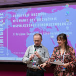 Kobieta i mężczyzna stoją. W tle ekran. Kobieta trzyma kartki i mikrofon, mężczyzna gra na instrumencie.
