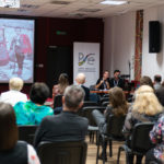 Kobieta i mężczyzna wygłaszają referat. Uczestnicy konferencji siedzą i słuchają. W tle na ekranie prezentacja multimedialna.