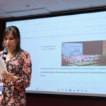Kobieta wygłasza referat. W rękach trzyma mikrofon i kartki. W tle na ekranie prezentacja multimedialna.