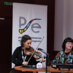 Dwie kobiety. Jedna wygłasza referat, druga gra na skrzypcach. W tle roll-up Polskiego Seminarium Etnomuzykologicznego.
