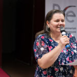 Kobieta trzyma mikrofon, kartkę i wygłasza referat.