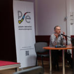 Mężczyzna wygłasza referat. W tle roll-up Polskiego Seminarium Etnomuzykologicznego.