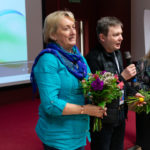Grupa trzech osób trzyma bukiety kwiatów w ręku. Mężczyzna mówi do mikrofonu.