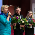 Grupa trzech osób trzyma bukiety kwiatów w ręku. Jedna z kobiet mówi do mikrofonu.