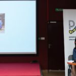 Dwie siedzące kobiety. Jedna z nich wygłasza referat. Na ekranie prezentacja multimedialna.