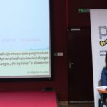 Dwie siedzące kobiety. Jedna z nich wygłasza referat. Na ekranie prezentacja multimedialna.