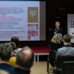 Dwóch mężczyzn prezentuje działalność ICTM. Uczestnicy siedzą i słuchają.