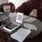 Stół z materiałami dla uczestników konferencji. Na stole Pismo Folkowe, zakładki, identyfikatory. W tle plecaki Polskiego Seminarium Etnomuzykologicznego.