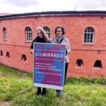 Dwie uśmiechnięte kobiety trzymają duży oprawiony plakat konferencji.