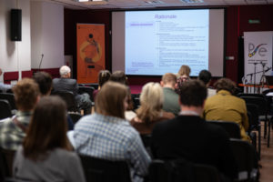 Uczestnicy konferencji siedzą na widowni, w tle wyświetlana prezentacja referatu