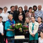 Grupa uśmiechniętych osób. W środku stoją trzy osoby trzymające wiązanki kwiatów. Przed nimi tort.